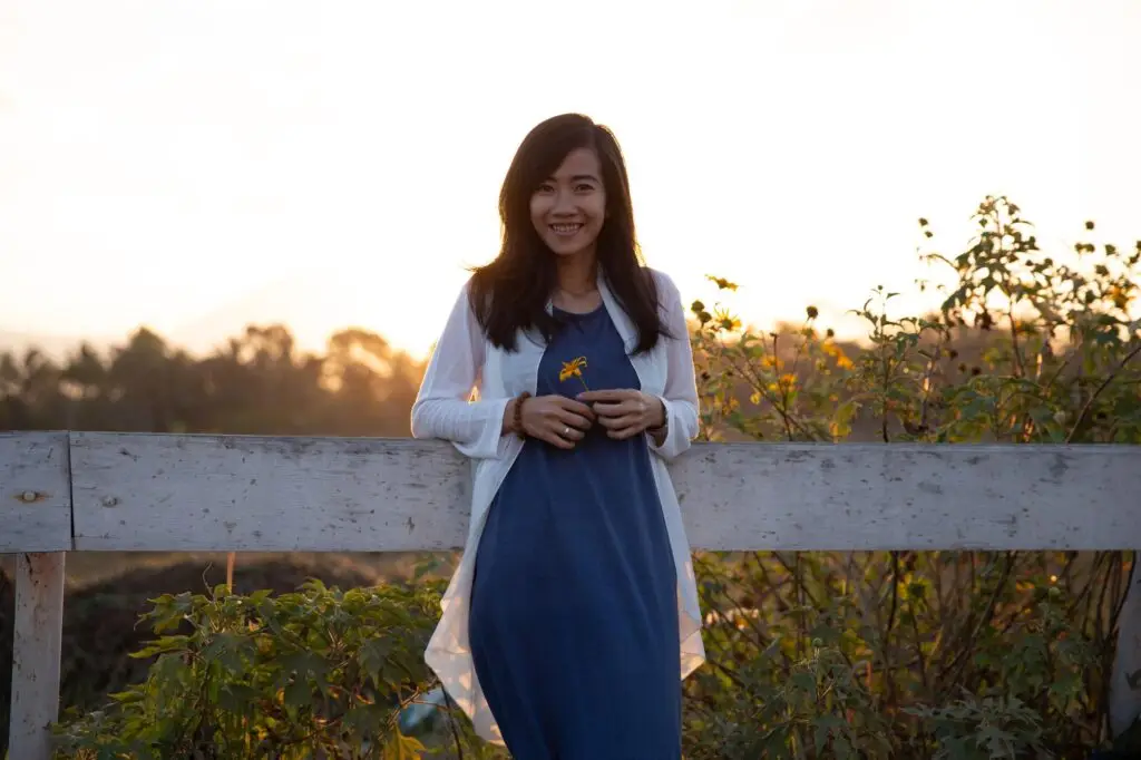 Smiling woman holding a flower, leaning on a white fence at sunset in a garden setting.