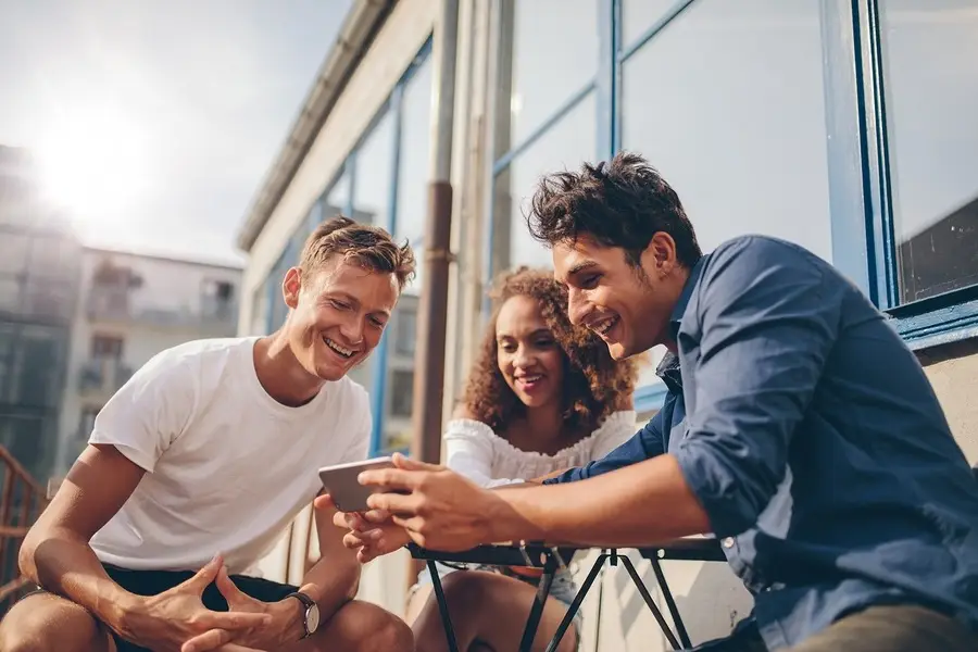 Three young friends smiling together outdoors, looking at a phone in early recovery and supportive connection.