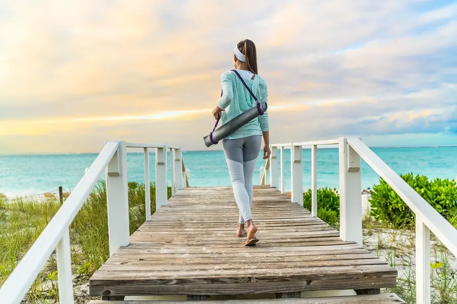 Woman carrying a yoga mat walks toward the ocean on a beach boardwalk at sunset, embracing holistic wellness.