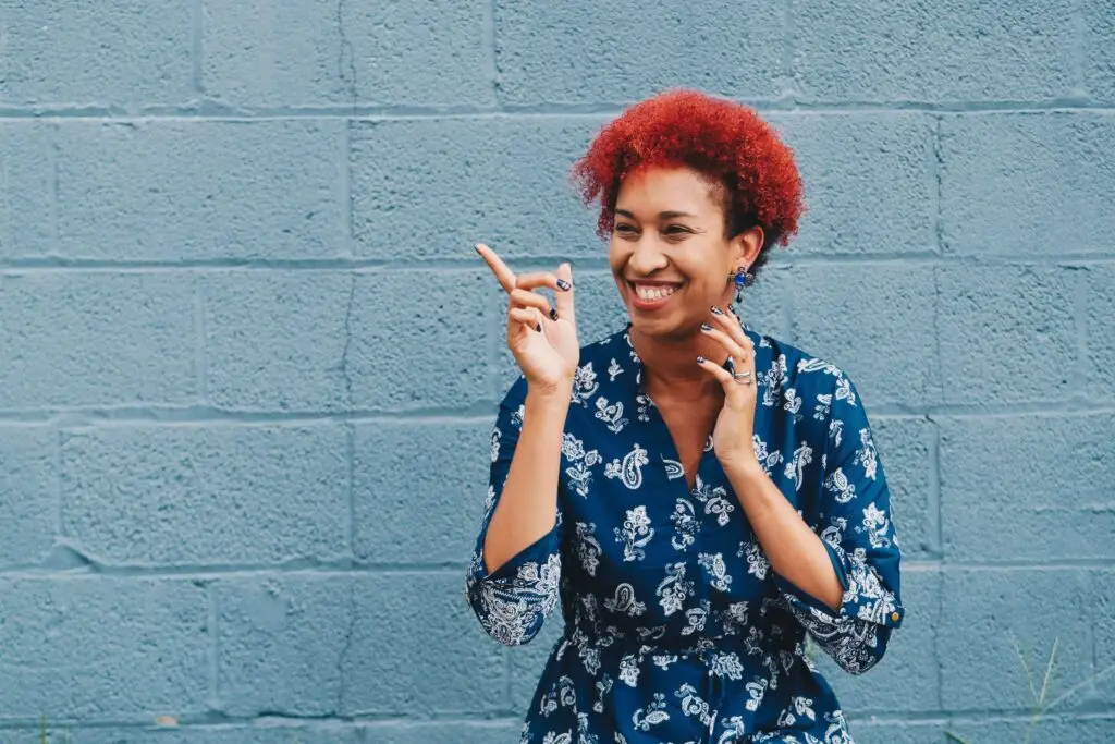 Smiling woman with red curly hair in a blue patterned dress standing against a blue brick wall.