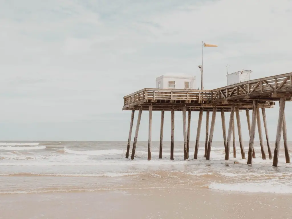 A weathered wooden fishing pier extending over gentle ocean waves under a soft, cloudy sky.