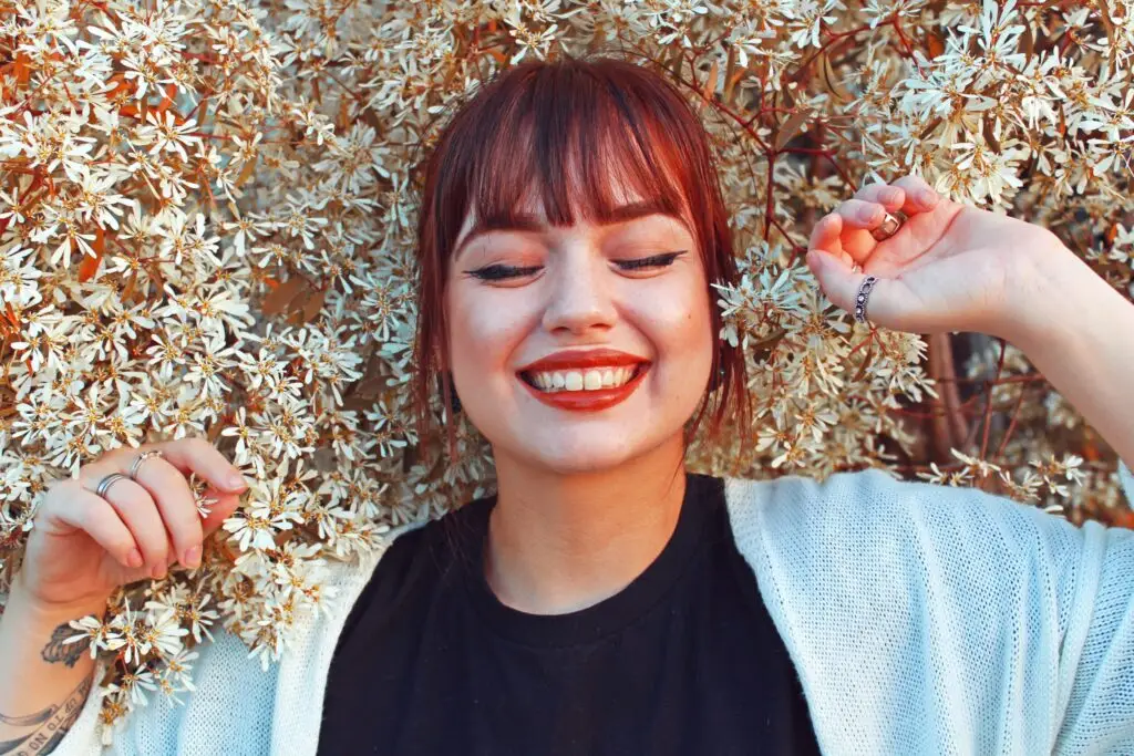 Smiling young woman with red hair and eyes closed, surrounded by white flowers, feeling joyful and at peace.