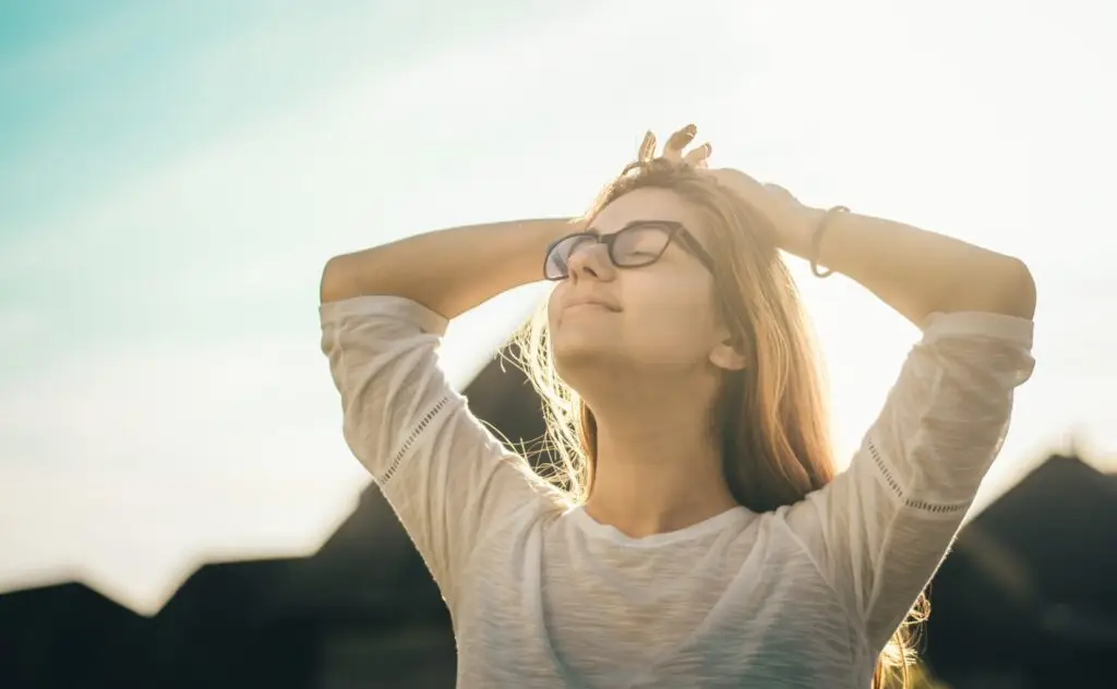Woman with eyes closed and hands in her hair, basking in sunlight, expressing peace and freedom in recovery.