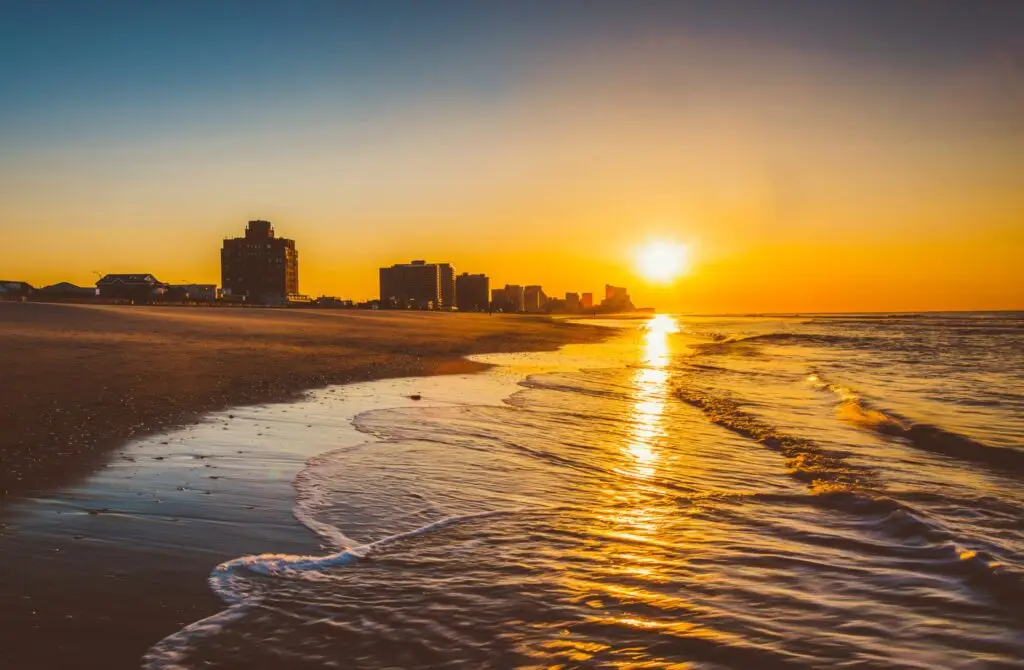 Golden sunrise over a calm beach with gentle waves and a distant city skyline silhouetted against the horizon.