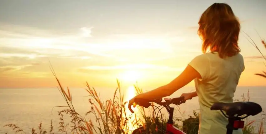 Woman with a bicycle gazing at a golden sunset over the ocean, symbolizing hope and a new beginning in recovery.